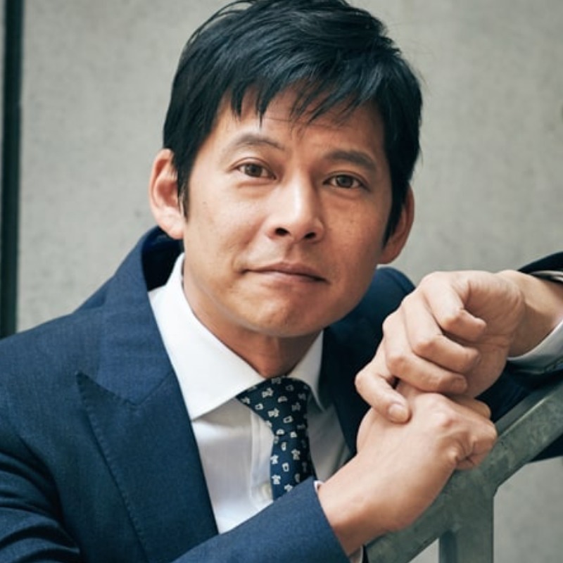 織田裕二が紺色のスーツで階段の手すりにもたれ掛かる画像