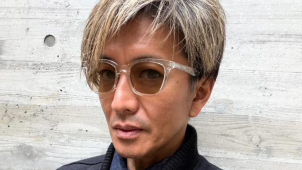 金髪ハイライトの短髪で、クリアフレームに黄色いレンズのサングラスをかけ、黒いジャケットを着た木村拓哉のクローズアップ写真。背景はコンクリートの壁。