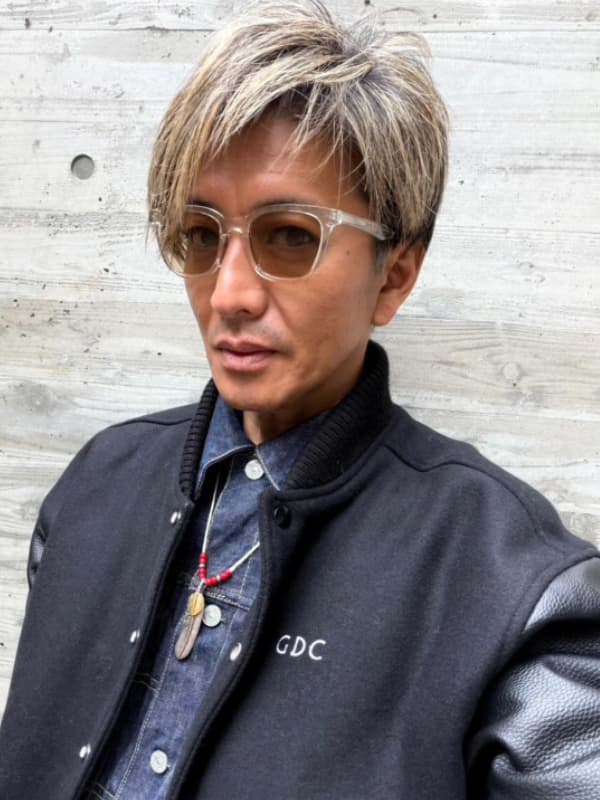 クリアフレームのサングラス、GDCのスタジャン、デニムシャツを着用し、コンクリート壁の前でポーズをとる木村拓哉の自撮り。