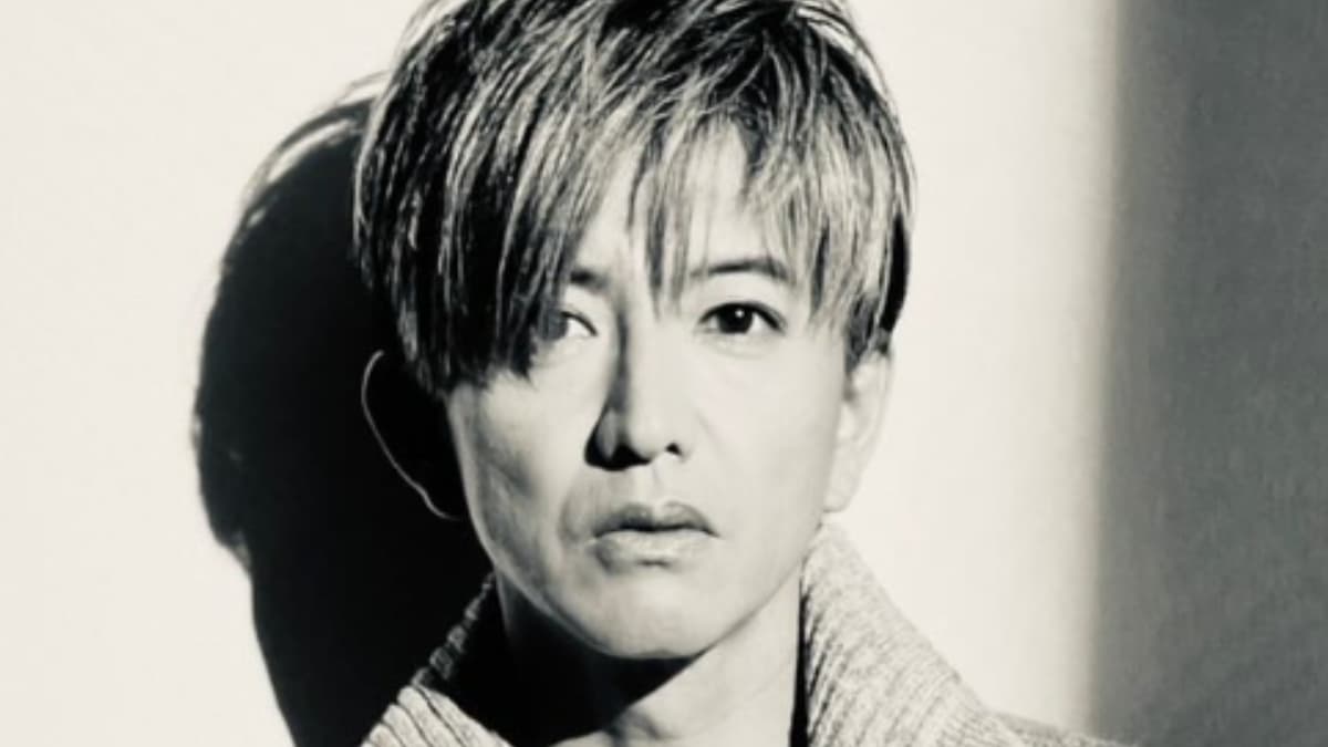 木村拓哉のモノクロクローズアップポートレート。強い眼差し、アッシュブロンドの髪、そして彼の影が強く落ちた明るい壁。
