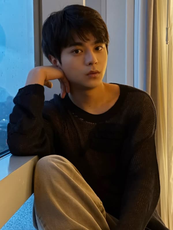 窓辺で頬杖をつき、リラックスした様子でこちらを見つめる佐藤勝利。透け感のある黒のメッシュニットを着こなしたポートレート。