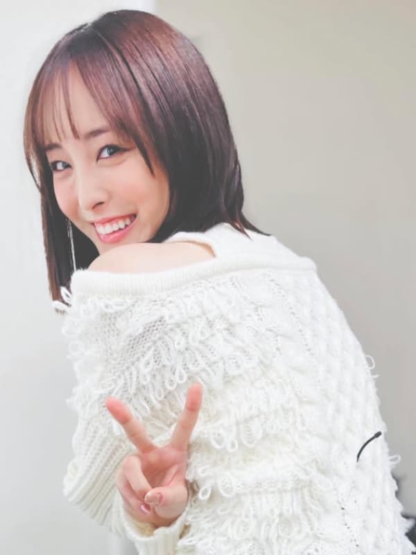 白いざっくりとしたニットを着用し、肩越しに振り返りながら満面の笑みでピースサインをする大友花恋。