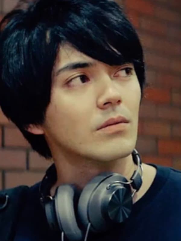 首にヘッドフォンをかけ、横を向いて何かを見つめる俳優の林遣都。暗い色のシャツを着用している。