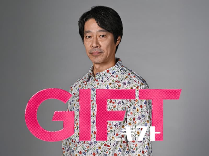 色鮮やかな花柄のシャツを着た俳優・堤真一のショット。画像手前にはピンク色の大きな文字でドラマタイトル「GIFT」および「ギフト」のロゴが配置されている。