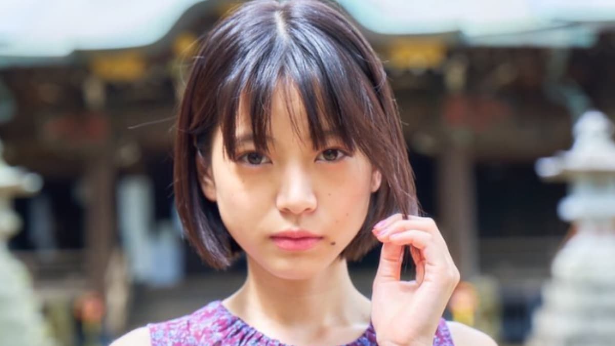 中井友望のポートレート。ボブヘアで、紫色の柄物ノースリーブトップスを着用。右手で髪を耳にかけるような仕草をし、カメラをまっすぐ見つめている。背景はぼかされた日本の神社のような建物と石灯籠。左目の下に泣きぼくろが確認できる。