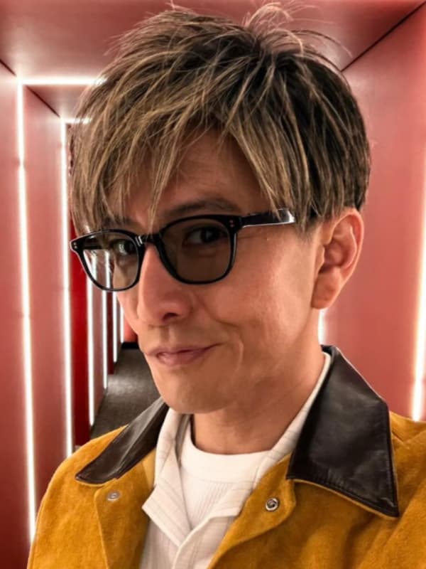 金髪ハイライトの短髪で、黒いフレームのサングラスをかけ、黄色のスエードジャケットと白いインナーを着た木村拓哉のクローズアップ写真。背景はライトアップされた赤い壁。