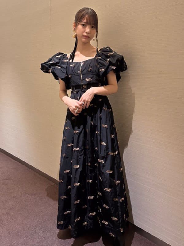 中井友望の全身ポートレート。髪を後ろでまとめ、黒のロングドレス(ゴールドの刺繍、パフスリーブ、フロントジッパー、ベルト付き)を着用。両手を体の前で交差させ、カメラを見ている。背景はぼかされたベージュの壁。