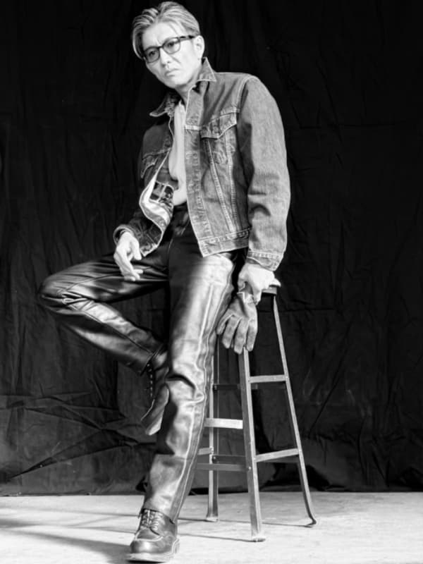 デニムジャケット、レザーパンツ、ブーツを着用し、サングラスをかけてスツールに座ってポーズをとる木村拓哉のモノクロ全身写真。背景は黒い布。