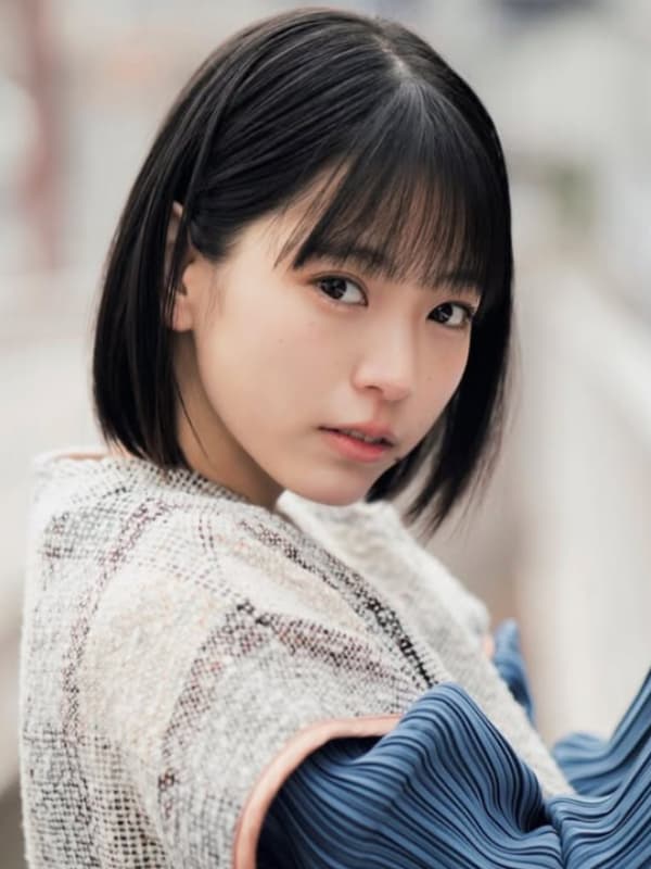 中井友望のポートレート。ボブヘアで、白とベージュのツイードジャケットと青いプリーツ袖の服を着用。少し斜めに構え、上目遣いでカメラをじっと見つめている。背景はぼかされた屋外。