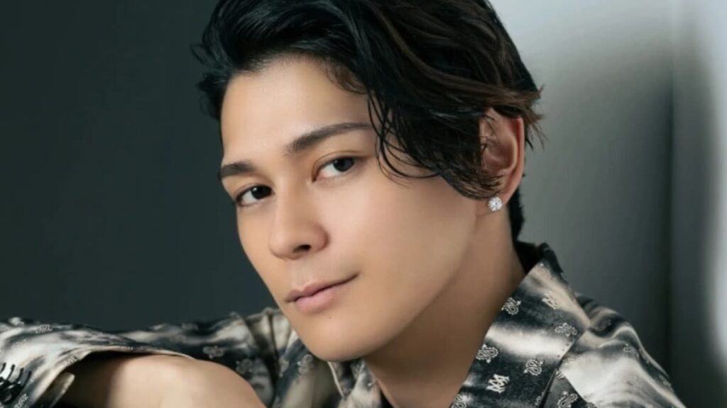 カメラを真っ直ぐに見つめる眞栄田郷敦さんのクローズアップ。白地に黒のペイズリー柄とロゴが施されたスタイリッシュな開襟シャツを着用。ウェット感のあるヘアスタイリングで、左耳には一粒のダイヤモンドピアスが光っている。