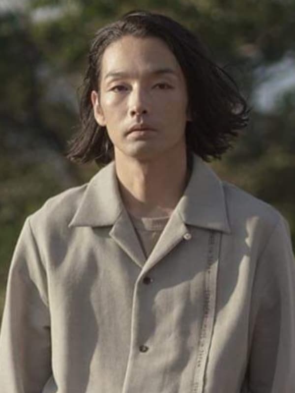 自然光の中で、風になびく中長髪と薄グレーのジャケット姿で物憂げに視線を落とす森山未來の屋外ポートレート。背景には緑豊かな森が見える。