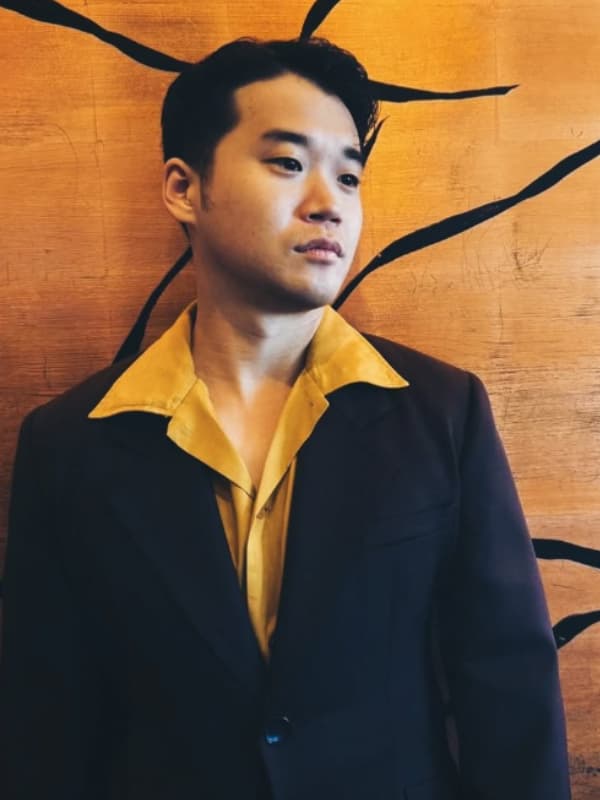 矢本悠馬氏が、暗い紫色のスーツに黄色のシャツを合わせ、木目調の背景に立つ。ミディアムショット。