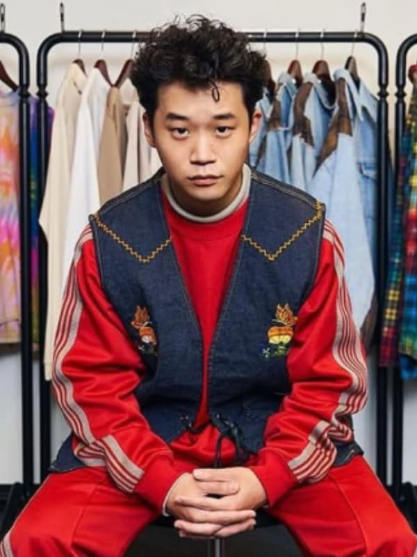 矢本悠馬氏が、赤地にストライプの入ったトラックスーツの上に、刺繍入りのデニムベストを重ね着して座る。フルショット。