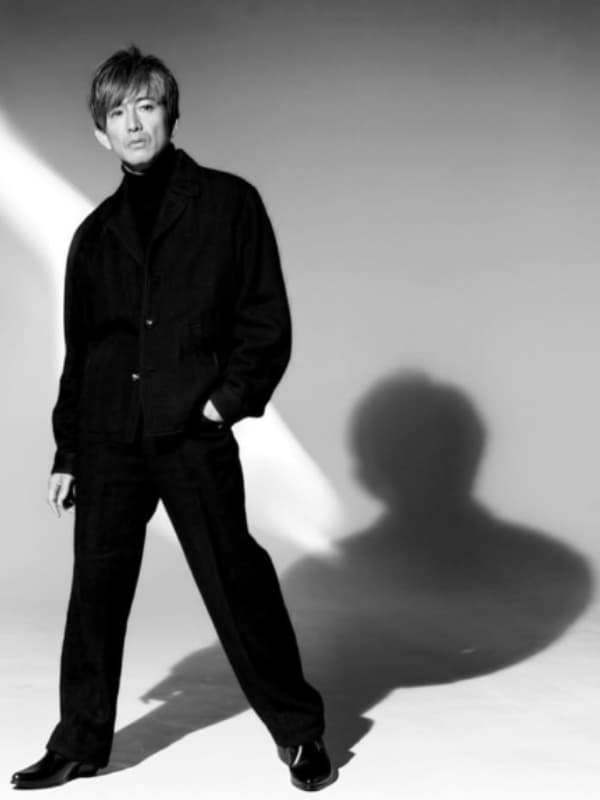 スタジオでポーズをとる木村拓哉のモノクロ全身写真。黒い服を着用し、背景の光と自身の大きな影が特徴。