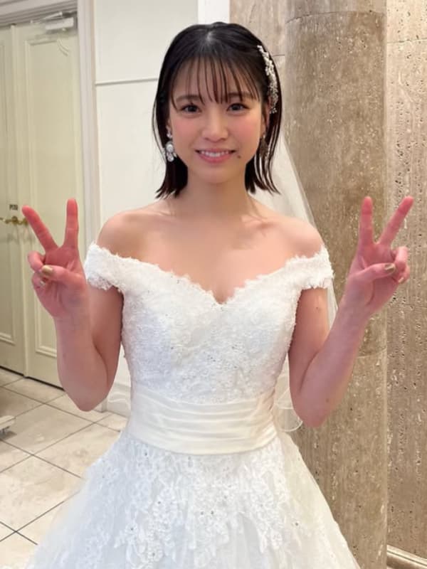 中井友望のポートレート。ボブヘアで、白のレースとオフショルダーのウエディングドレス(サテンベルト付き)を着用。正面を向き、両手でピースサインを作り、笑顔でカメラを見ている。背景は大理石の壁とドア。