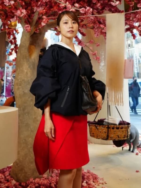 ボリューム袖が特徴的なネイビーのトップスと鮮やかな赤のスカートを纏い、黒いバッグを持つ有村架純。背景には桜の装飾があり、華やかで都会的なファッションショット。