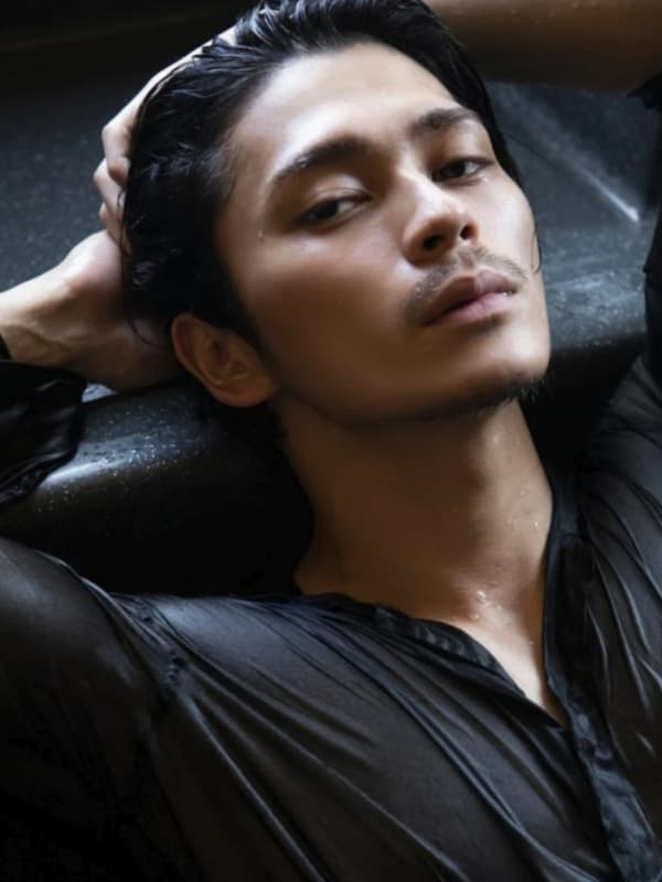 濡れた透け感のある黒いシャツを纏い、両手を頭の後ろに置いて横たわる眞栄田郷敦さんの官能的なショット。顔や首筋、シャツに水滴がついており、ミステリアスでセクシーな雰囲気を醸し出している。