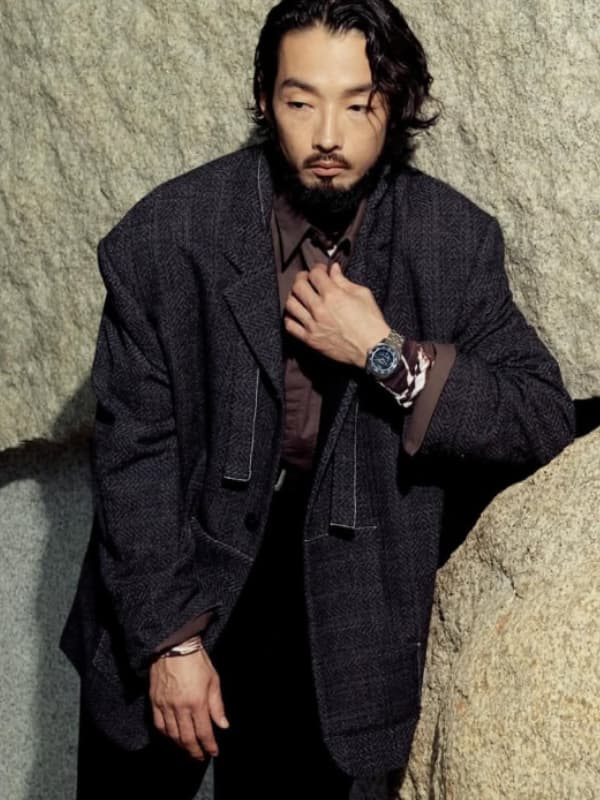 荒々しい石壁に背をもたれかけ、髭を生やした姿でダークなツイードジャケットの襟を直す森山未來。左腕には腕時計を着用しており、ドラマチックな照明に照らされている。