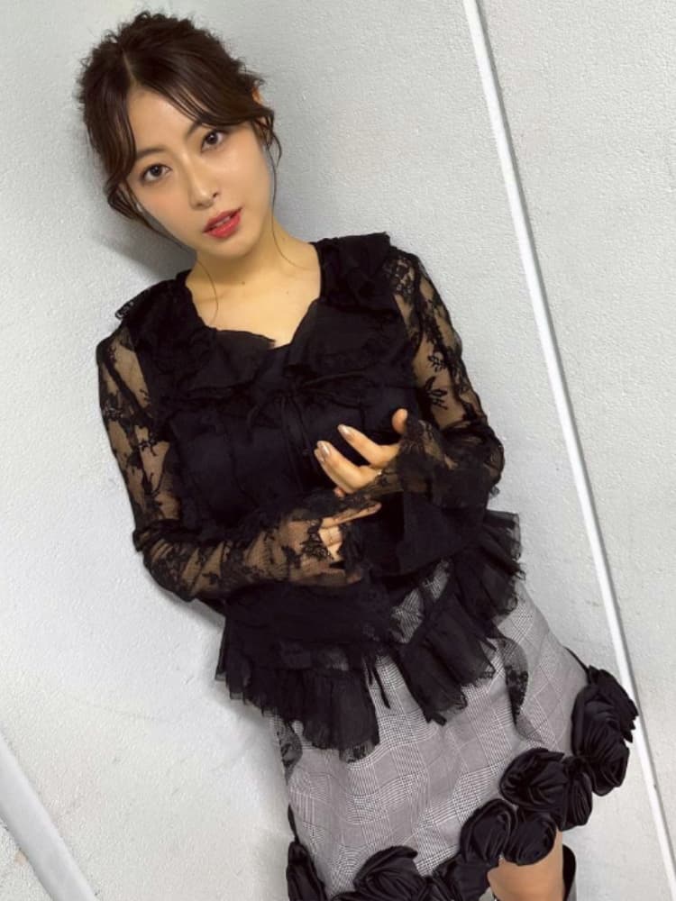 壁に寄りかかり、カメラを見る瀧本美織氏。黒いレースのブラウスとグレーのチェック柄のスカートを着用。