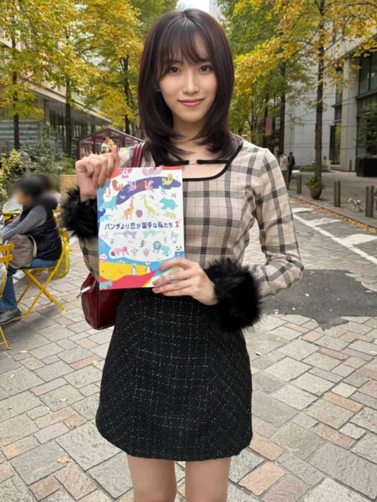 街角に立つ、茶色のショートヘアの片岡凜。チェック柄の長袖ブラウス(胸元にスリットあり、袖口に黒のファー付き)と黒のスカートを着用。手に「パンダより恋が苦手な私たち 1」と書かれた本を持って、微笑んでいる。