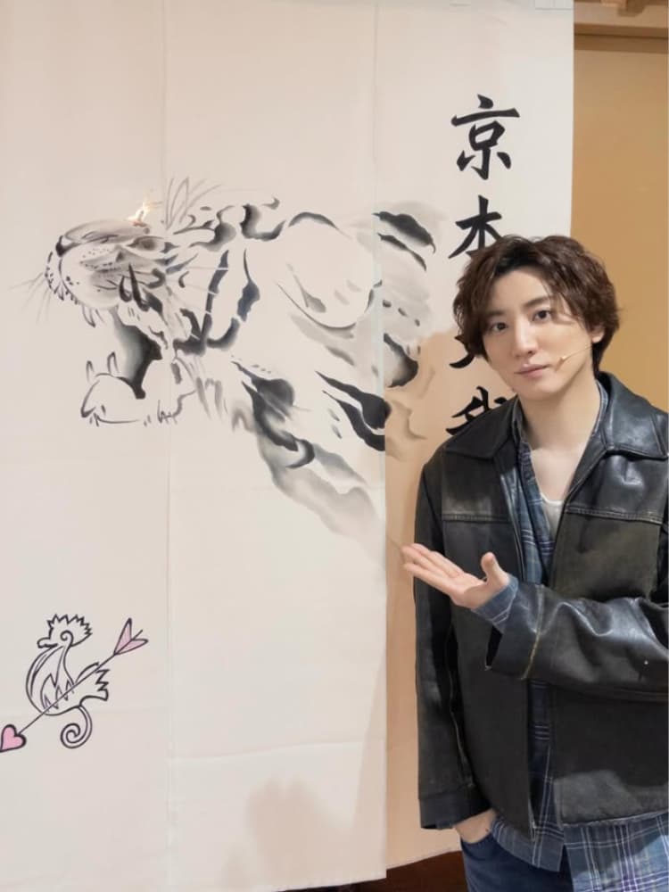 黒いレザージャケットと青いチェックシャツを着用した京本大我が、咆哮する虎の水墨画、達筆な書（「京本」の文字）、ピンクのハートが付いた矢の絵がある和風の展示物を右手で差し出している。
