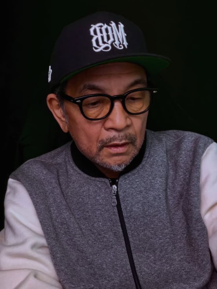 眼鏡とグレーと白のジャケット、白いロゴが入った黒い野球帽（つばのウラが緑）を着用した中野英雄氏が、暗い緑色の背景で、何かを下向きに見つめているアップ写真。