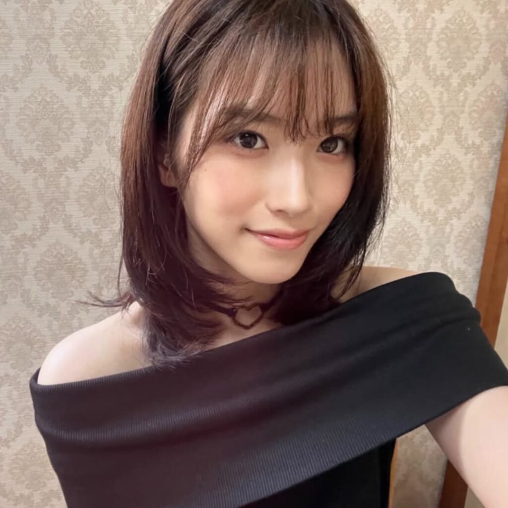 オフショルダーの黒いトップスを着た、茶色のショートヘアの片岡凜の自撮り。彼女はカメラに向かって優しく微笑んでおり、背景は模様のある壁紙。