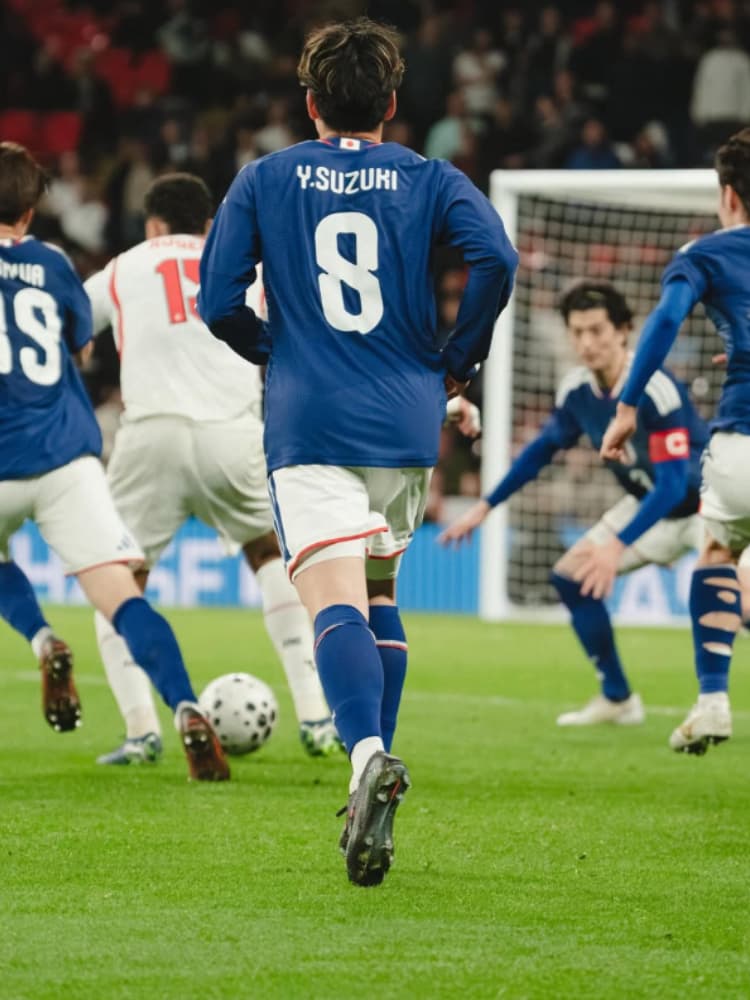 サッカー日本代表の青いユニフォームを身に纏い、ゴール方向へ走る鈴木唯人選手の後ろ姿。背番号8と「Y.SUZUKI」のネームが鮮明に写っている。