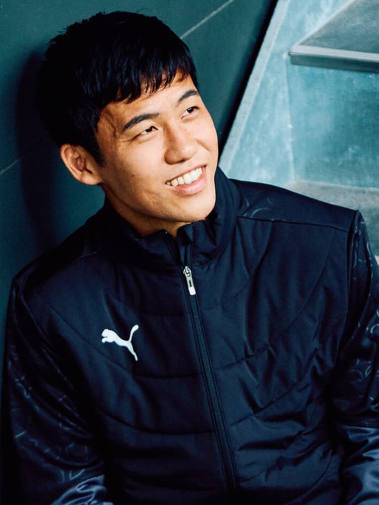 黒いプーマのジャケットを着て、階段に座り壁に背を預けながら、リラックスした表情で斜め上を見て微笑む遠藤航選手。