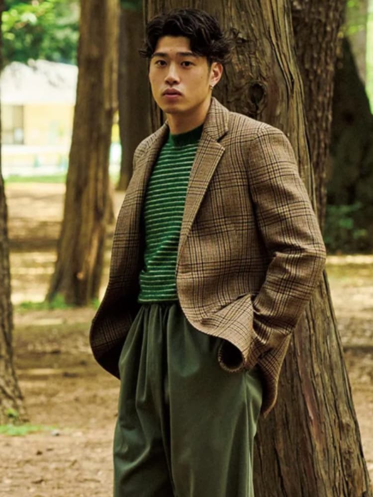 森の中のような背景で、茶色のチェック柄ジャケットと緑のボーダーニットをスタイリッシュに着こなす、モデルのような佇まいの鈴木唯人選手。