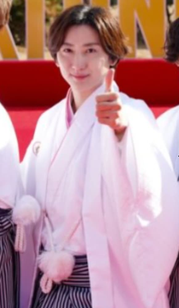 白い羽織、ピンクの着物、縦縞の袴を着用した京本大我が、紅白幕の前で、右手の親指を立ててサムズアップをしている。