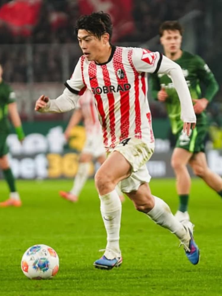 スタジアムの照明の下、集中した表情でボールを運びながら相手ディフェンダーを抜き去ろうとするSCフライブルクの鈴木唯人選手。