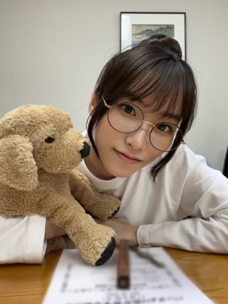 室内でテーブルに座る、茶色のショートヘアで眼鏡をかけた片岡凜。白の長袖Tシャツを着て、茶色の犬のぬいぐるみを抱きしめている。テーブルの上には書類がある。