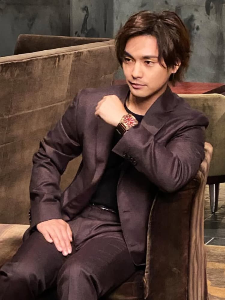 ブラウンの光沢感あるスーツを纏い、赤い文字盤が印象的な高級腕時計をアピールするように椅子に座る柳楽優弥。大人の色気が漂うポートレート。