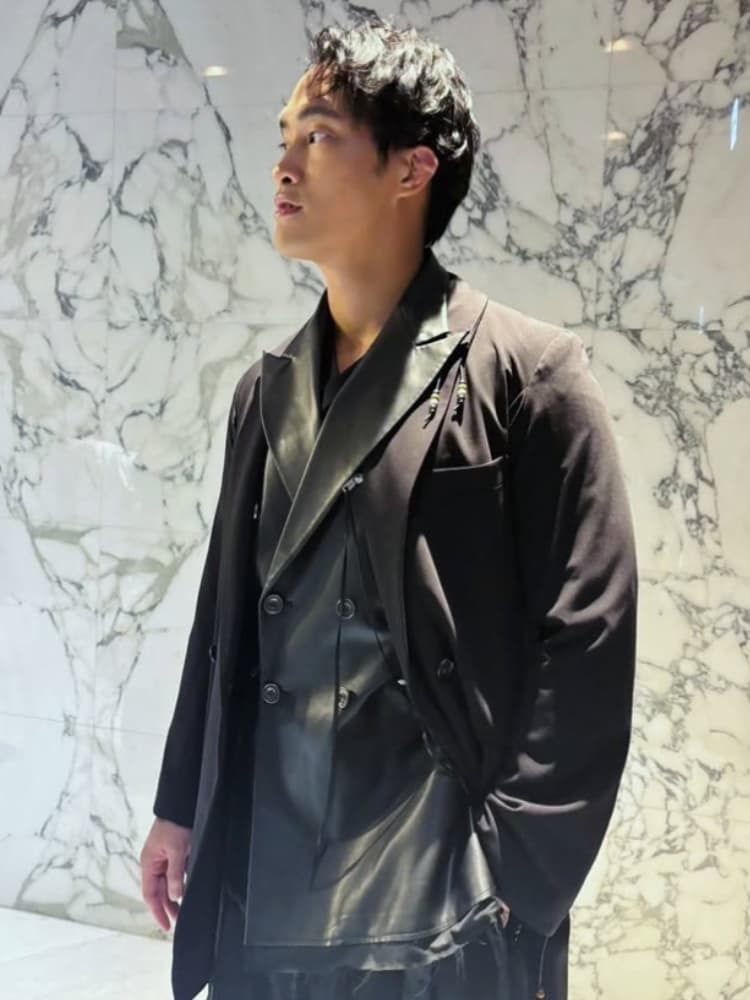 大理石の壁の前でポーズをとる佳久創。黒いスタイリッシュなコートとレイヤードジャケット、レザーのディテールが特徴的な衣装を着用し、斜め上を見つめている。俳優としての洗練された雰囲気と、190cmを超える身長、筋肉質の体格を活かした衣装映えするシルエット。『Tarzan』でのインタビューや現代劇における「強者」としての存在感を放つ。