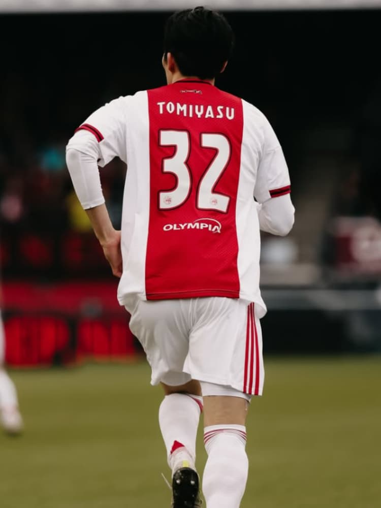 アヤックスの2025-26シーズンホームキット（赤と白、背番号32、ネームTOMIYASU）を着用し、ピッチ上を走る冨安健洋の背面写真。