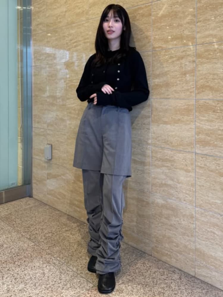 大理石風の壁に寄りかかり、黒いカーディガンとグレーのパンツを着て立つ白石聖の全身ショット。