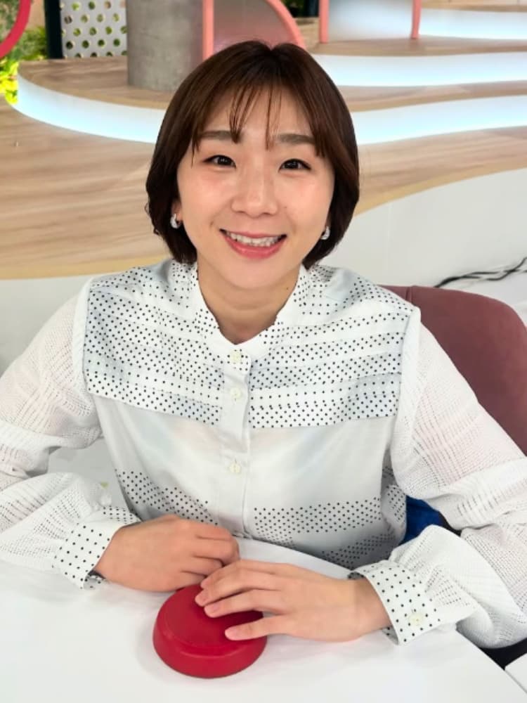 白いドット柄のブラウス姿で、赤いボタンが置かれた白いテーブルに座って微笑む高橋成美さんの写真。