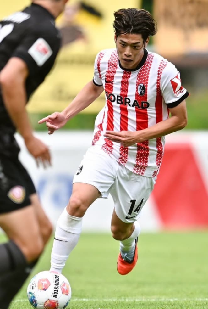 SCフライブルクの白と赤の縦縞ユニフォームを着用し、真剣な表情でドリブルを仕掛ける鈴木唯人選手。躍動感あふれるプレーシーン。