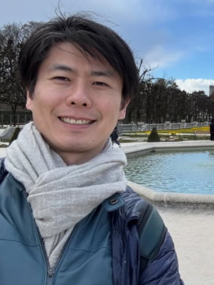 ヨーロッパの庭園の噴水前で、マフラーを巻いて微笑む福間洸太朗のセルフィー。