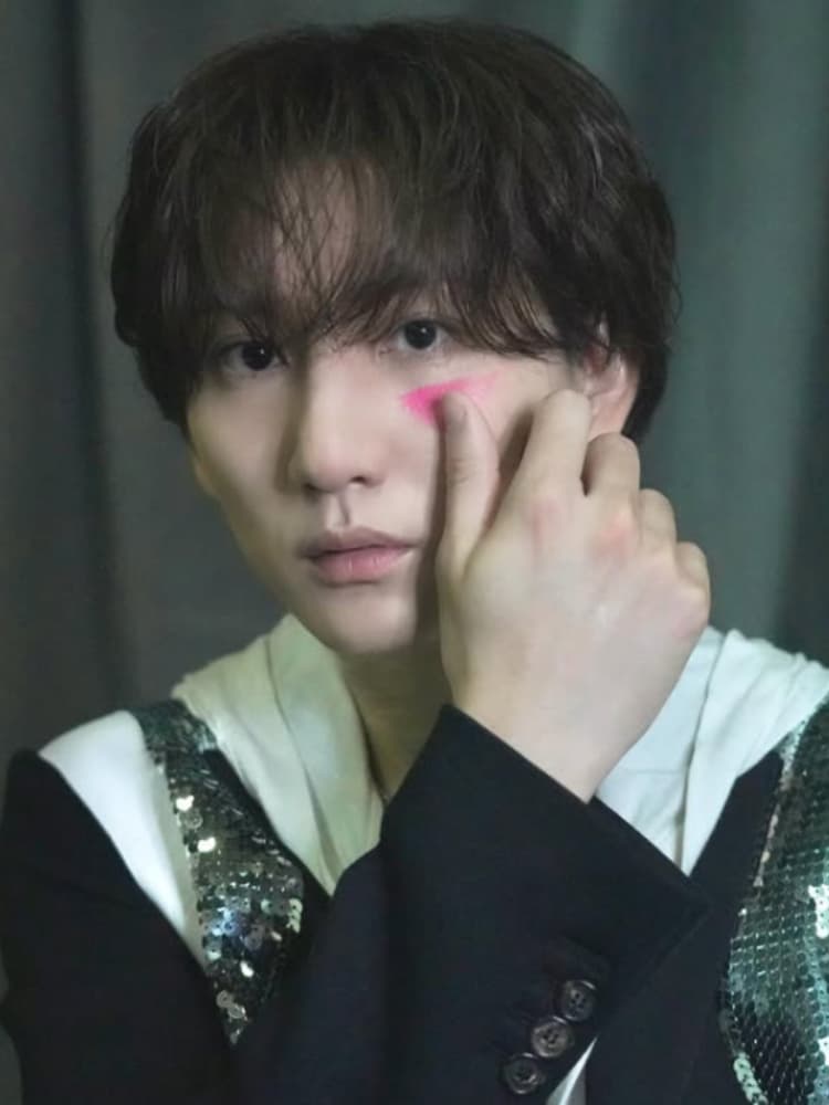 松村北斗氏。黒い髪が少しラフにスタイリングされている。カメラをじっと見つめている。真剣な表情。黒いジャケットの下に、白い襟付きシャツ、さらにその下に、黒いタートルネックを着用。左の頬に、ピンク色のチーク（またはペイント）が指で塗られたような跡がある。右手を頬に当て、人差し指でピンク色のチークを指している。