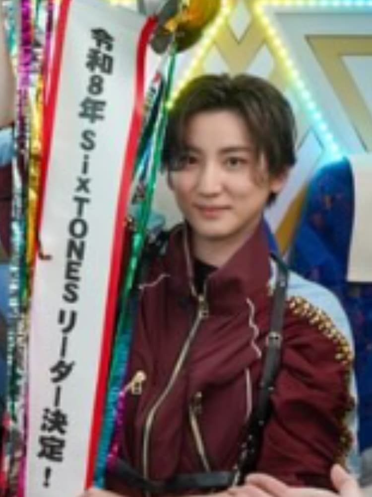 ボルドー色のジャケットと白いアクセントのある衣装を着た京本大我が、ステージで「令和8年 SIXTONES リーダー決定！」と書かれた垂れ幕を持っている。黄色と緑の照明。