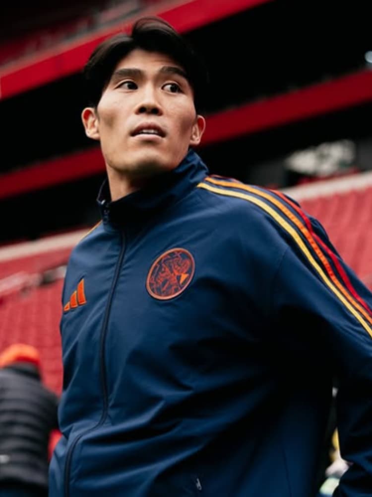 ヨハン・クライフ・アレナの観客席を背景に、アヤックスのネイビーブルーのトレーニングジャケットを着用して上を見上げる冨安健洋。