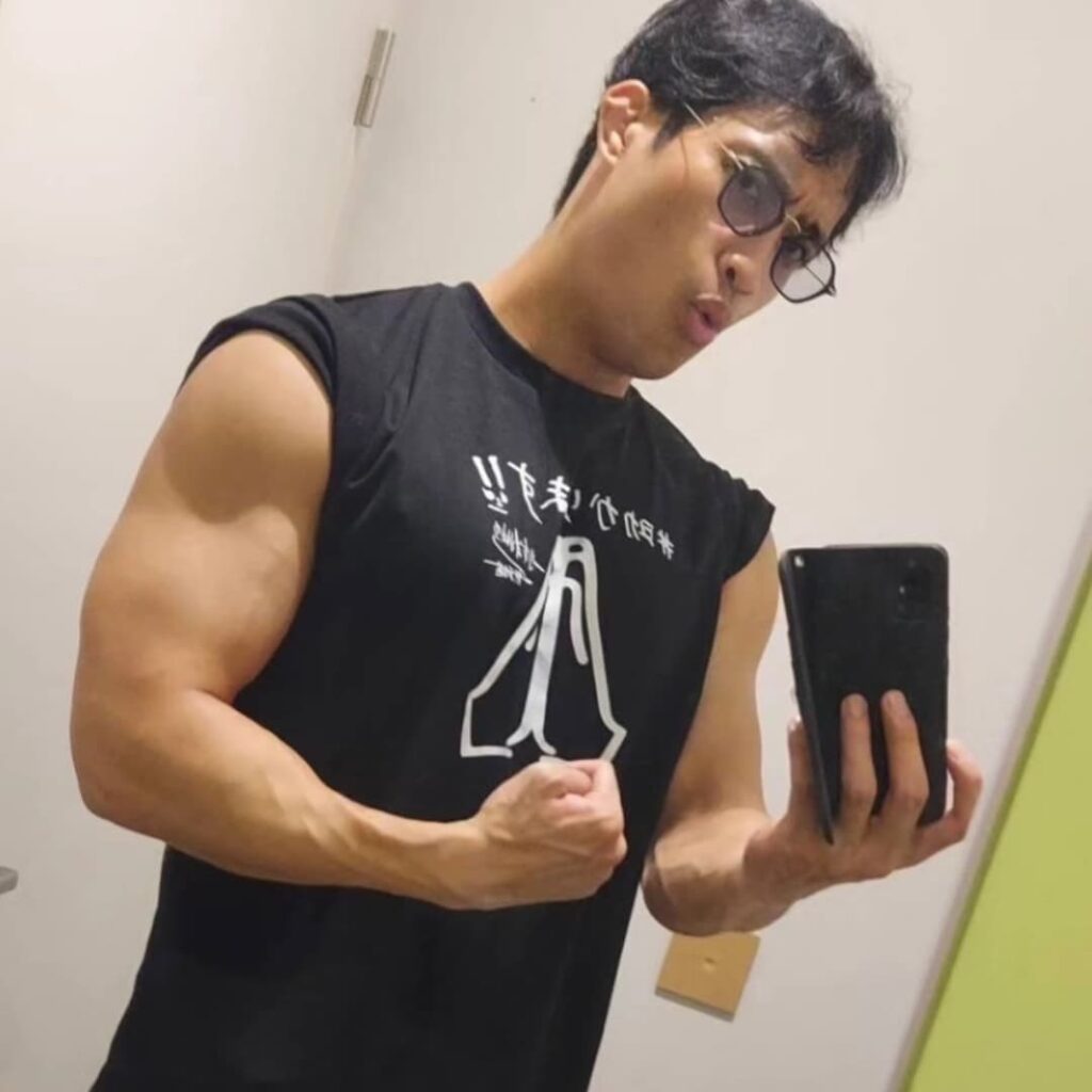 鏡越しのセルフィーを撮る佳久創。サングラスをかけ、ノースリーブの黒いTシャツから発達した上腕二頭筋を誇示している。Tシャツには、自身が演じたNetflixシリーズ『サンクチュアリ -聖域-』の力士役「龍谷龍貴」を思わせる文字と、四股を踏む力士のイラストが描かれている。日々の鍛錬で培われた圧倒的な肉体美を象徴する一枚。