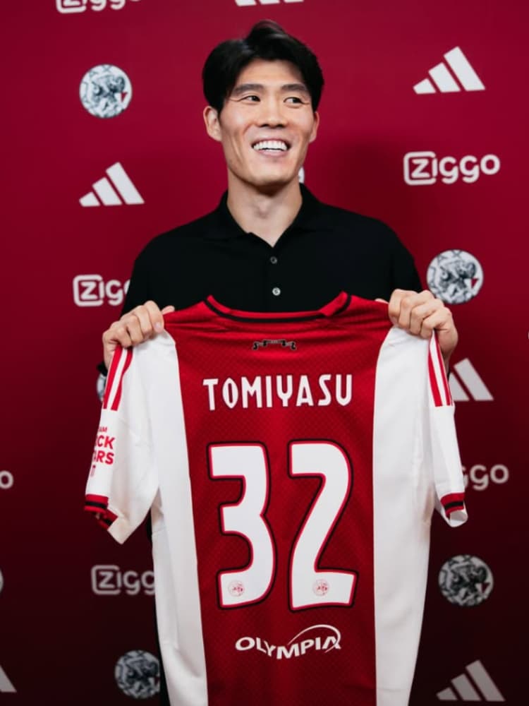 アヤックスの入団発表にて、スポンサーロゴの背景の前で笑顔で背番号32の「TOMIYASU」ネーム入りホームキットを掲げる冨安健洋。