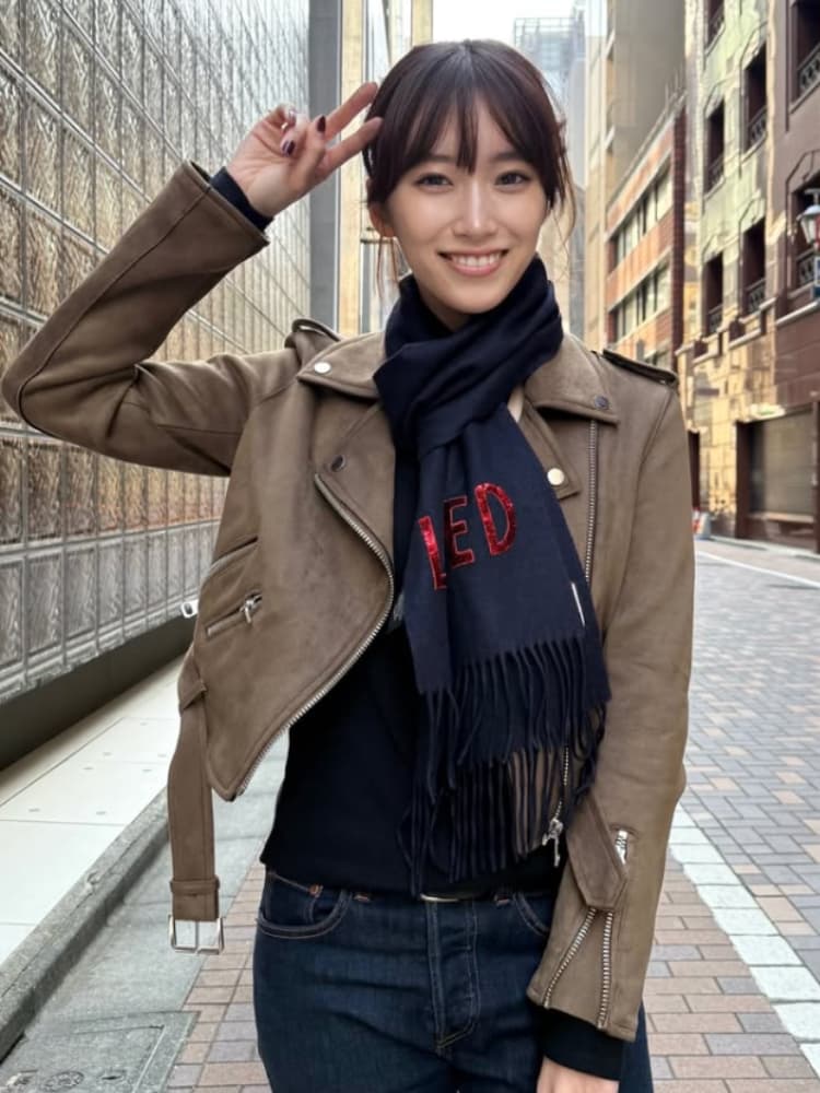 街路に立つ、茶色のショートヘアの片岡凜。茶色のレザージャケット、黒いニット、ネイビーのスカーフを着用し、スカーフには赤字で「LOVED」のロゴがある。カメラに向かってピースサインをし、笑顔を見せている。