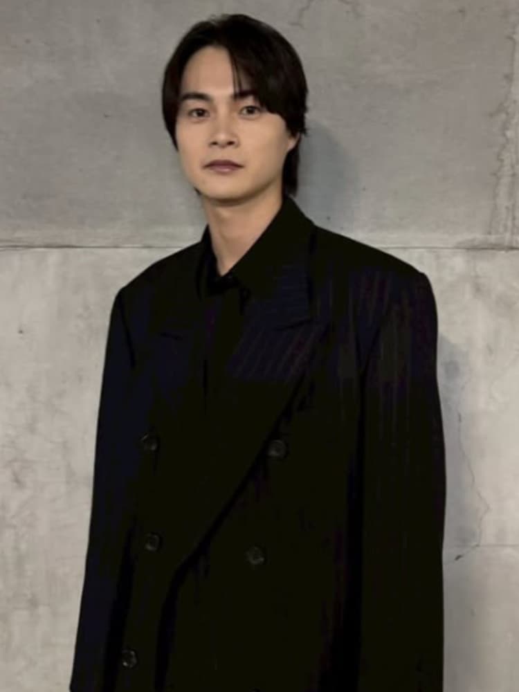 コンクリート壁の前でポーズをとる俳優の結木滉星氏の全身カット。黒いセットアップ姿。