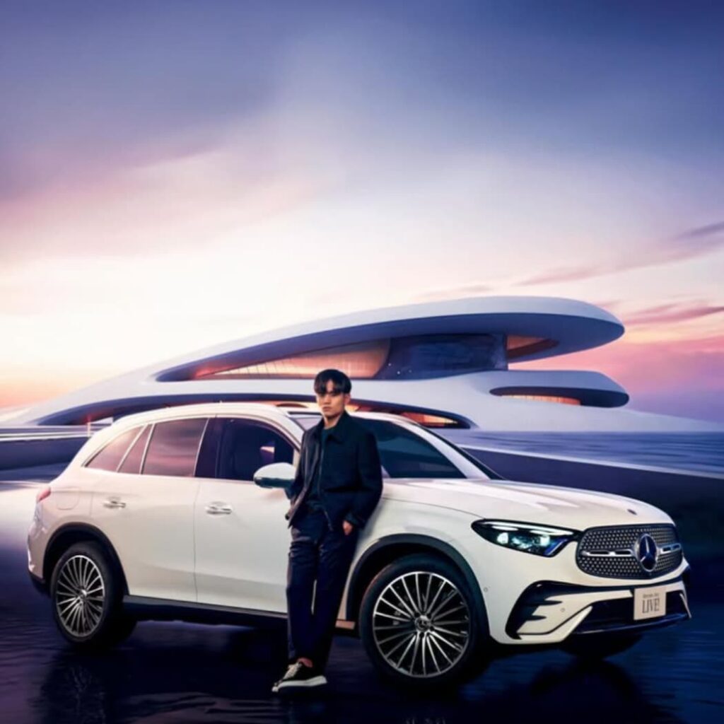 未来的な建物を背景に、白いメルセデス・ベンツGLC SUVに寄りかかるカジュアルな服装の久保建英の全身ショット。