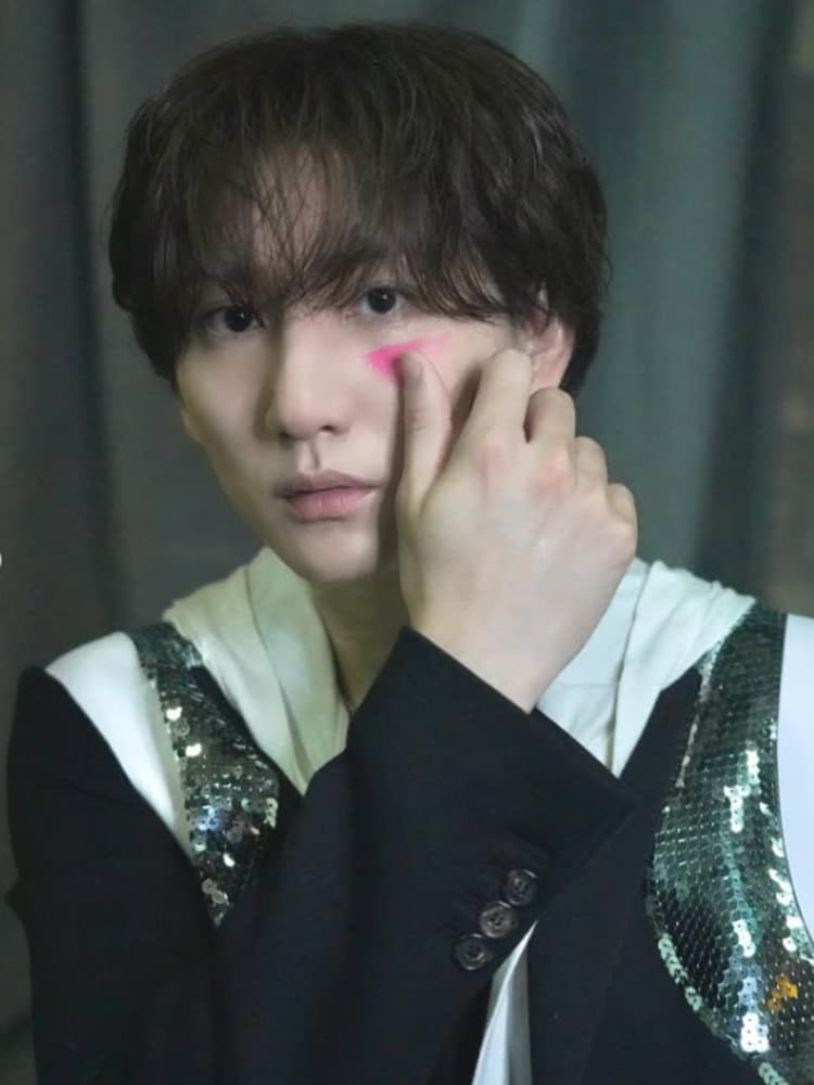 黒いジャケットと白いインナーを着用した京本大我。左頬にあるピンク色のメイクアップに左手で触れている。グレーのカーテンを背景に、正面を見つめている。