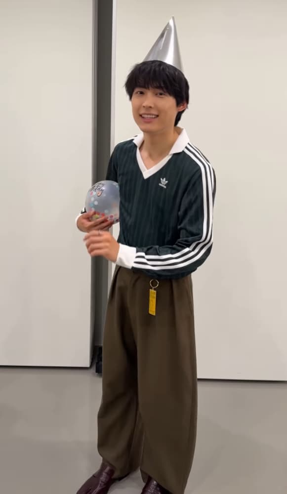 松村北斗氏。黒い髪。少し微笑んでおり、親しみやすい表情。濃いグリーンのVネックのスポーツシャツ（アディダスのロゴと白い3本線あり）の下に、白いTシャツを着用。全身ショット。左手に、白い小さな風船をいくつか持っている。頭には、シルバーのパーティーハットを被っている。背景は白い壁。